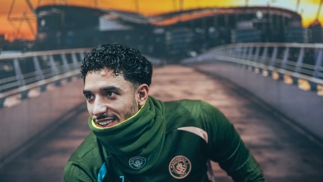 A Manchester City player in training gear with a blurred face, the background shows the Etihad Stadium during sunset.