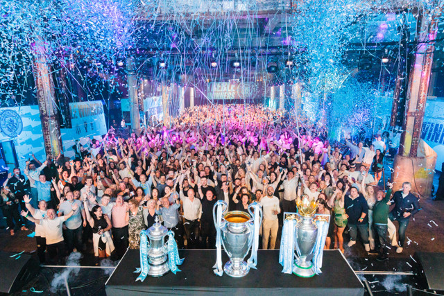 Large crowd celebrating in an indoor venue with three trophies displayed in the foreground surrounded by confetti and streamers.